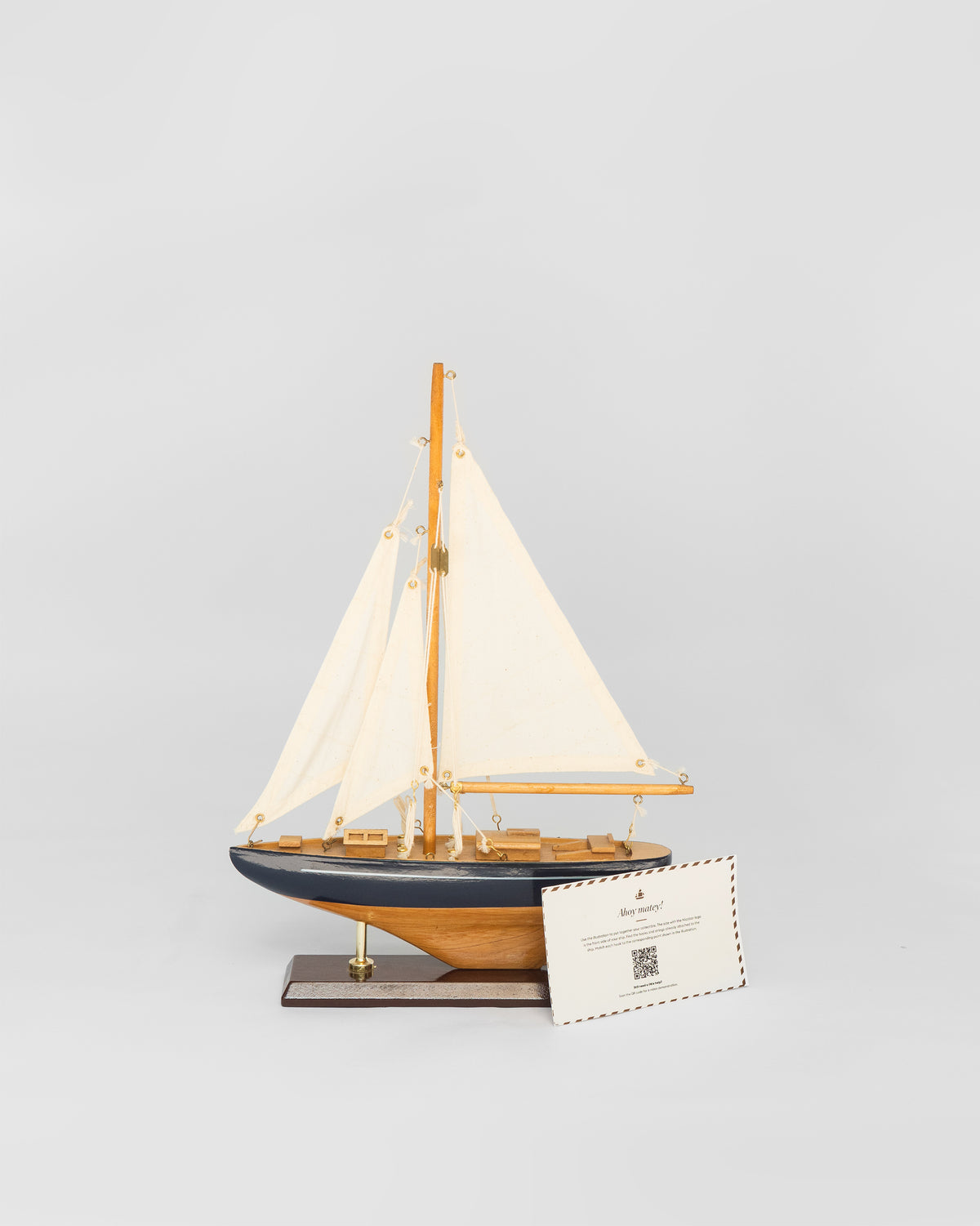 Small Sail Boat Home Accent, a wooden model with cream sails and a navy hull, with a card beside it.