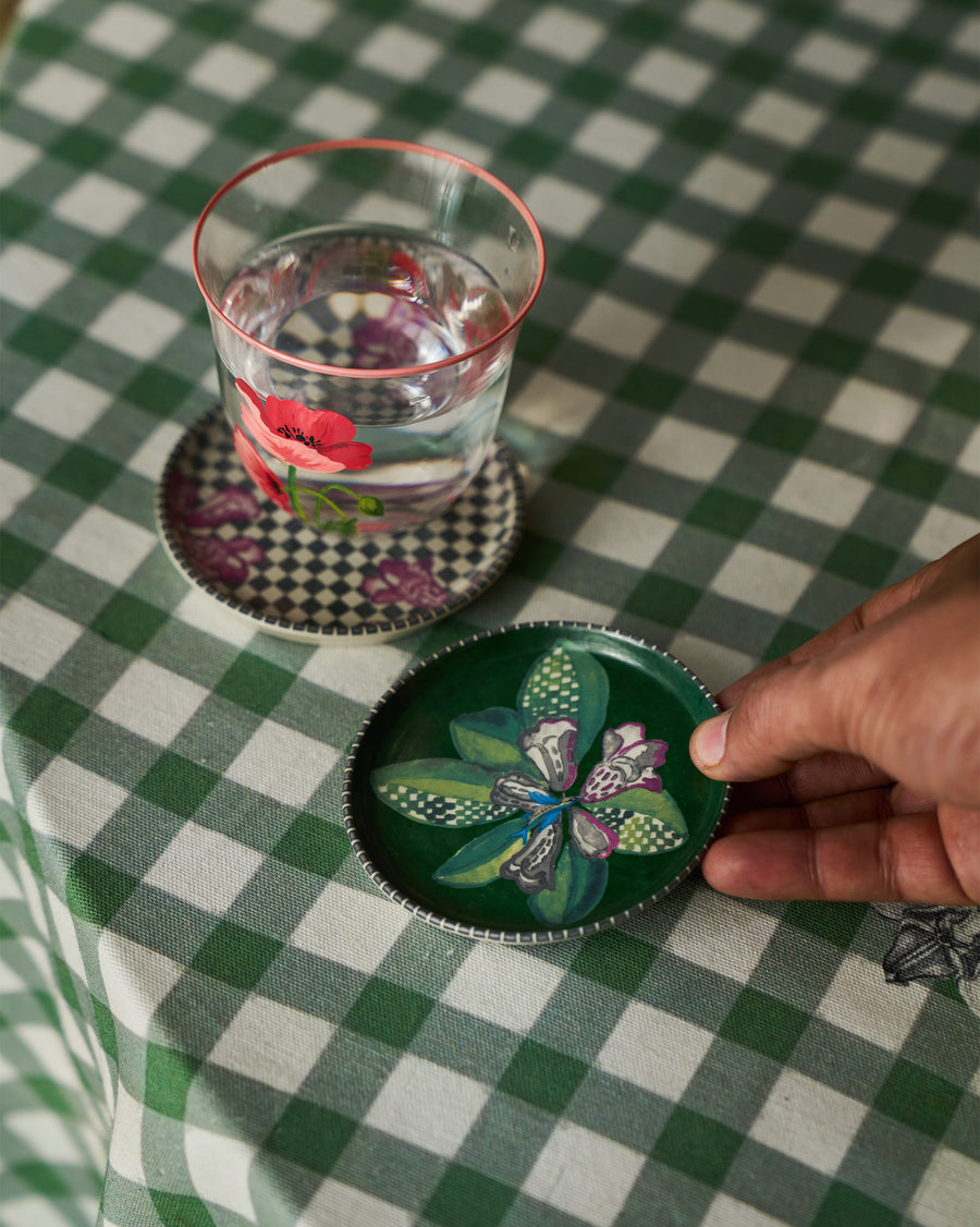 A hand placing a green Reesha coaster beside a glass on green gingham