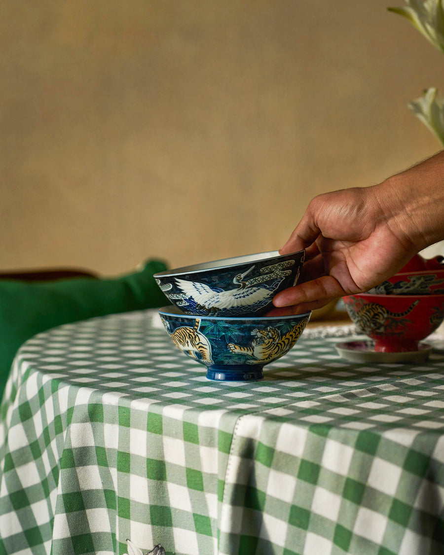 A hand stacking blue Crane and Tiger bowls on a green gingham table