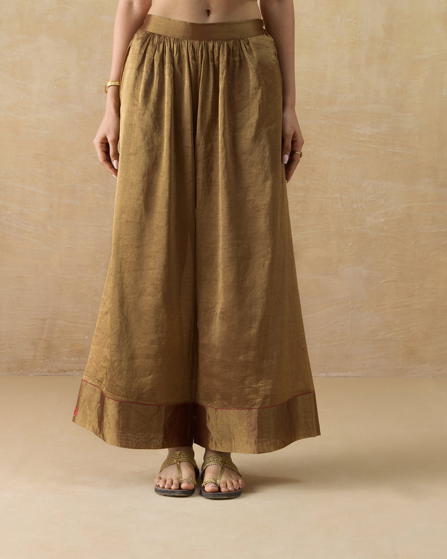 The Charisma Pants, wide-leg gold metallic tissue pants with a deep hem band, worn with flat sandals.