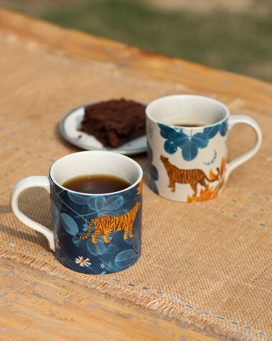 A navy Nadi mug of coffee and a cream one beside a brownie on jute