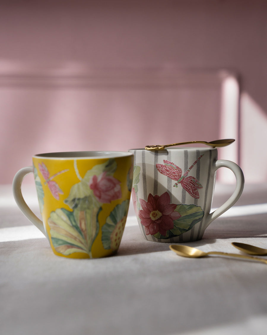 A yellow and a striped Lotus Garden mug with gold spoons against a pink wall